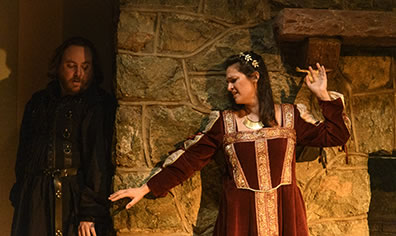 Production photo by Kenneth Garrett of Ophelia, leaning against the flagstone wall of the fireplace, reaching out her hand toward Hamlet hiding in a nook. 