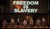 The cast stand and crouch next to chairs, shouting, except Winston sitting, and O'Brien standing at the back in front of the office canteen door; overhead is a video image of the Brotherhood leader with "Freedom Is Slavery" in bold white letters sumperimposed over him