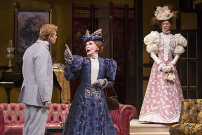 In a richly appointed apartment of red leather and gold brocade, Lady Bracknell in a purple embroidered hat, stiff colar, fancy hat, and gloves points a finger at Algernon in gray suit as Gwendolen in puffy-shouldered, pink print dress and floral hat watches.