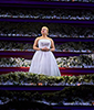 Under a spotlight, Eva Peron wearing a bared-shoulder, white ball gown, sings amid layers of candlit fwhite flowers.