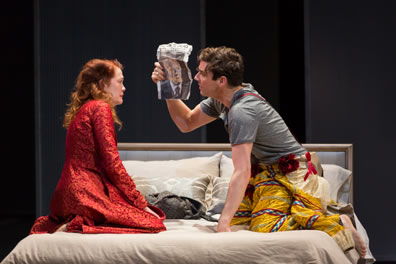 Hamlet in gray t-shirt and jester's pants holds up a small poster of his father to Gertrude, wearing a red robe, both kneeling on the bed.