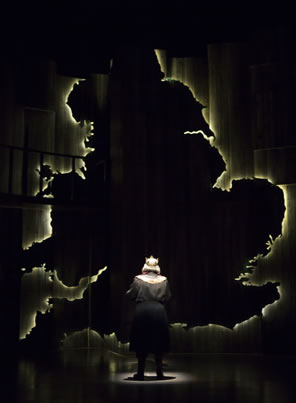 Henry, back to us, stands in a spotlight looking up at a giant silhouette map of England outlined in white light.