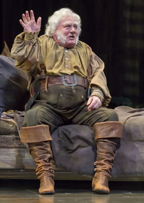 Falstaff in dirty yellow shirt, striped green pants and brown boots waves his right hand as he sits on a bed.