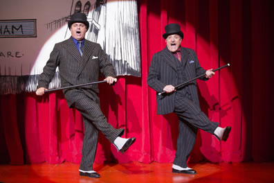 The two mobsters in classic mobster dark gray pinstripe suits but wearing tophats and holding canes kick out their right legs as they sing in a spotlight against a red curtain