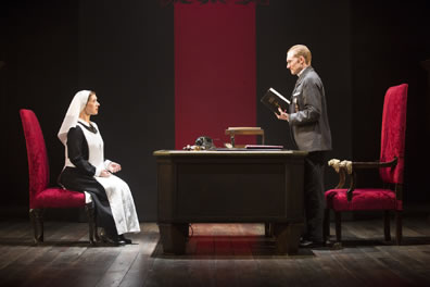 Isabella sits in a red chair across the desk from Angelo, standing, holding a Bible, large throne-like red chair behind him, and a red banner hanging down int he background