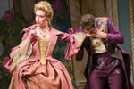 Lucille in a pink cloak over a pink floral-patterend hoop skirt with yellow lace corset and drooping lace sleeves, her blond hair tied up in curls, Dorante in purple pants and long coat with psychodelic splotches on the pants and coat trim, and a yellow shirt. He is kissing her left hand, she's looking the other way.