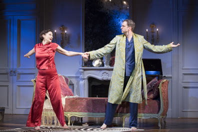 Amanda in red pajamas and Elyot in blue pajamas with green print robe dance with arms outstretched, holding hands, in front of a chez lounge in an apartment with marble mantled fireplace, large mirror, and wall candelabra