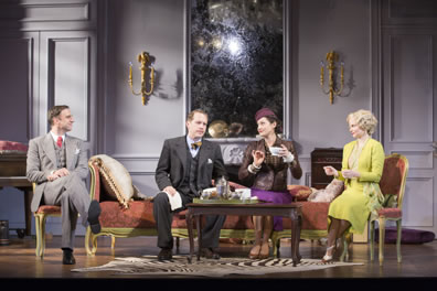 Elyot in a gray three-piece suit with red tie and handkerchief sits crossed-leg in a chair and looking smugly over at the chez lounge on which Victor in dark gray three-piece suit, bowtie and handkerchief with napkin in hand, Amanda in purple dress and hat and gold-embroidered jacket and teacup in hand, while Sibyl in yellow dress sits in a chair to the right. The tea set is on a table in front.