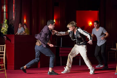 Tybalt in purple jacket, blue pants, no socks and black shoes thrusts a knife at and holds the right arm of Mercutio with a black and white running jacket, dirty white pants and tennis shoes, Romeo in the background with gray Tee and black pants, and around them is the red decor of the catering banquet hall.