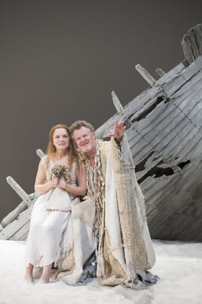 Miranda, rag doll clutched to her beast in a simple white shift dress, sits next to Prospero wearing a burlap cloak decorated in pieces of driftwood. He is pointing up and outward, both smiling, and in the background is the ruined bow of a boat sticking up from the white sand.
