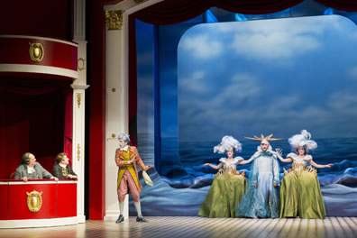 Dangle and Sneer sit in red theater box seats to the left, Puff stands near them talking to them, and to his left in the center of the stage are two women dressed as shorelie (green wide hoops skirts with sillhouettes of towns on the hips and clouds for hats) and a man dressed as an ocean with sun rays for a hat. Behind them is the English channel, a mechanism emulating waves. 