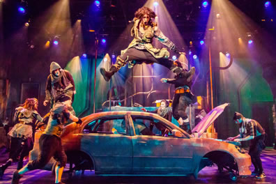 A guy with long hair and patchwork coat leaps, spread legged, high over an abandoned car as other dancers frolic and Amiens plays his keyboard in the opened hood of the car. Pipes are in the background against green and purple wall and multi-color lights.