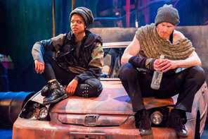Rosalind in black knit cap, camoflage shirt, black flack vest, and black pants with rivets sits on the hood of the car with Orlando in gray knit cap, shaw, and black pants, holding a bottle of booze.