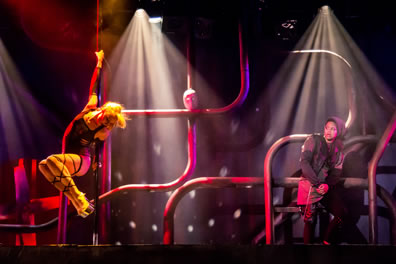 Phoebe to the left has her hands extended along a pole and her legs, with 5-inch platform shoes, tucked up as she dances for Rosalind, wearing a gray hoodie and chained to one of the serpentine pipes at the back of the stage; Jacues mask is sitting at an intersection of two poles in the center of the image.