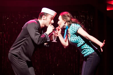 Benedick in a sailor uniform and Beatric in blue polkadot blouse and tight jeans sip through straws out of a single ice cream float glass.