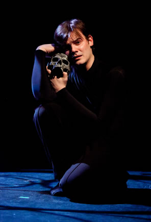 Hamlet all in black blending into black background squats on the foor holding a blackened skull up against his cheek