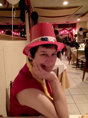 Sarah wearing a red sleeveless blouse, a plastic red party top hat, and a yellow lei with restaurant tables behind her.