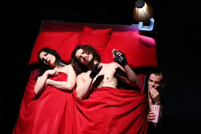 Kate and Petruchio in a bed with red sheets watching porn TV as Grumio, eating popcorn,  peers out from under the bed 