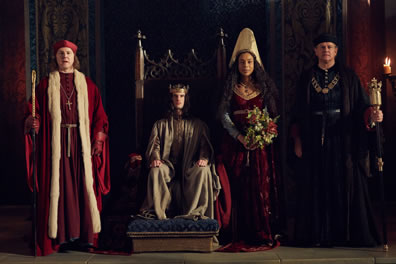 Winchester in log red robe with white fur trim, priest hat, scepter stands next to Henry in gold robe and crown sitting on throne, next to Margaret in red dress, gray cloke, white high hat and holding a bouquet of roses standing next to Gloucester in black robes and chain around his neck.