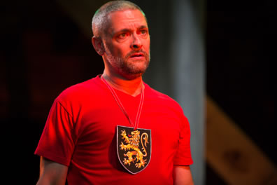 Talbot facing forward talking in soliloquy wears a red t-shirt with a royal lion crest hanging around his neck.