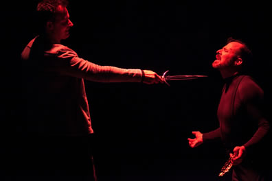 Hamlet stands with his knife pointed at the throat of the King who is kneeling, hands outstretched and laurel wreath in his left hand, his head tilted back as he prays to heaven. Both are cast in a red light on a dark background.