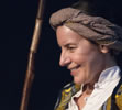 A grinning Prospero, gray cloth turban on his head, yellow cloak over white linen shirt, dark wood staff with section of light wood next to his face.