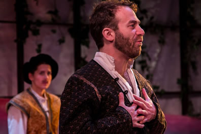 Orsino in black jacket with gold brocade, white shirt opened down the chest and his hands crossed on his chest; in the background, Viola is gold brocade vest, white shirt, and black renaissance cap.