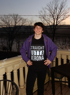 Meehan, wearing his Straight Outta Verona black t-shirt, black pants, and purple jacket stands with hands on hips on a balcony with the river in the background at dusk