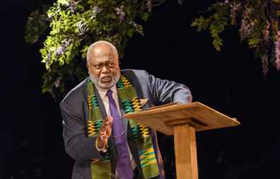 Preacher in gray suit, purple tie and an African scarf oer his shoulders stands at a podum with blooming tree branches overhead.