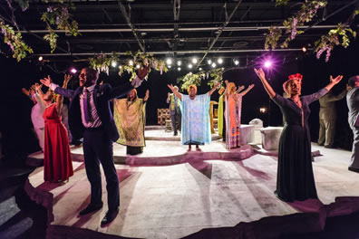 Singers around a three-step stage with their hands upraised, the light grid overhead.