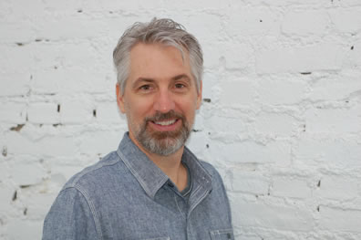 Gray hair, gray-and brown beard and mustach, denim shirt, smiling at the camera in front of a white-painted brick wall