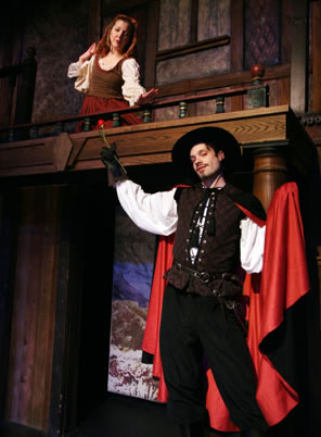 Henriquez on lower stage in cavalier brown lace-up vest, white blouse shirt, black cape with red lining and black wide-brimmed hat holding a rose in his right hand, while up in the balcony Violante holds out her hand coyly; she's dressed in simple olive green tank over white blouse and orange dress