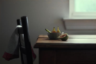 Two apricots in a bowl on a wood table, one apricot sliced in half next to them, a wood kitchen chair next to the table with a dish towel draped over the back, the window sill in the background and bright light coming through the window