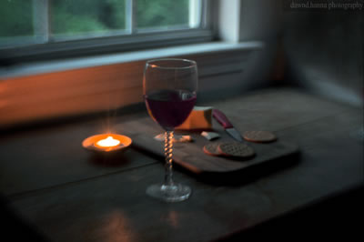 A glass of red wine on a counter next to a candle with a board of crackers and cheese behind